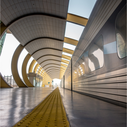 A modern train station with sleek architectural arches, reflective metal panels, and a bright pathway leading toward the platform’s exit.