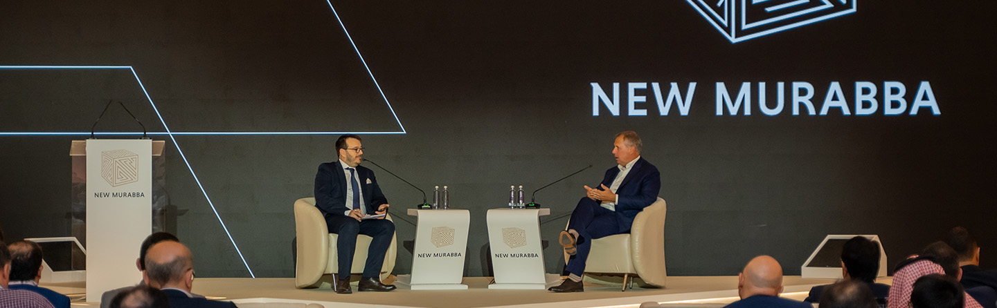 نقاش جماعي في منتدى شراكة نيو مربع، يجلس فيه رجلان على المسرح مع شعار نيو مربع في الخلفية.